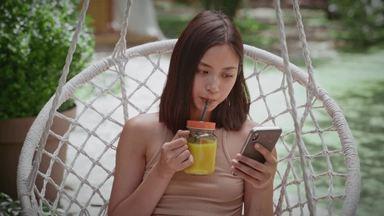 a woman sitting in a hanging chair and looking at her smartphone while drinking orange juice